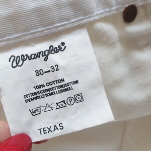 May include: Close-up of a white Wrangler jeans tag. The tag displays the brand name, size 30-32, and fabric composition: 100% cotton. Washing instructions and the word "TEXAS" are also visible.