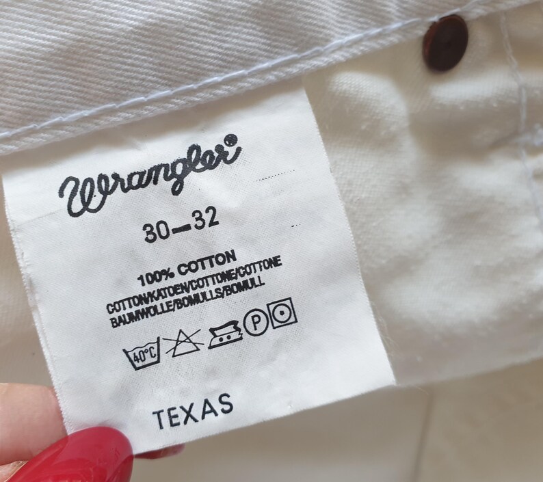 May include: Close-up of a white Wrangler jeans tag. The tag displays the brand name, size 30-32, and fabric composition: 100% cotton. Washing instructions and the word "TEXAS" are also visible.