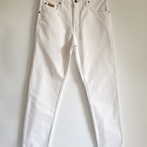 May include: A pair of white denim jeans hanging on a black hanger. The jeans have a classic five-pocket design with a button and zipper closure. A small brown patch is visible on the right front pocket.