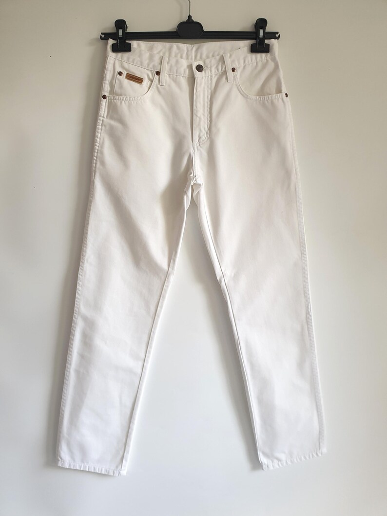 May include: A pair of white denim jeans hanging on a black hanger. The jeans have a classic five-pocket design with a button and zipper closure. A small brown patch is visible on the right front pocket.