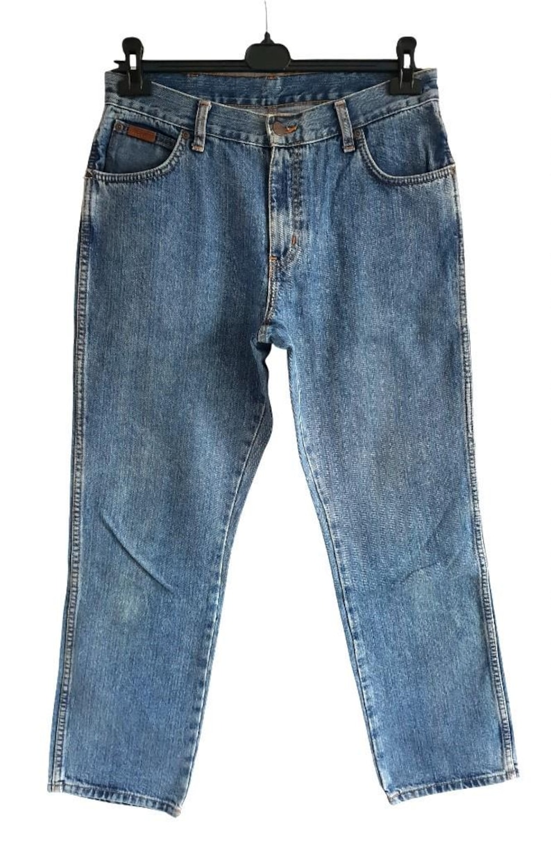 May include: Light blue denim jeans with a classic five-pocket design. The jeans are hanging on a black hanger against a white background. The jeans have a straight leg cut and a button and zipper closure.