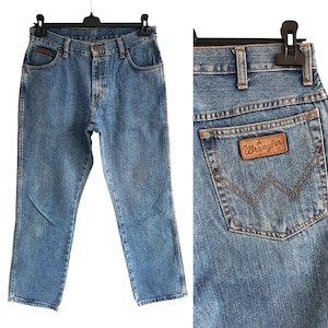 May include: Light blue denim jeans hanging on a black hanger. The jeans have a classic five-pocket design and a straight leg fit. The back pocket features a brown leather patch with the Wrangler logo.