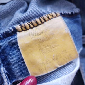May include: Close-up of a pair of blue denim jeans. The yellowed tag reads "Wrangler" with size information and care instructions. The jeans show signs of wear.