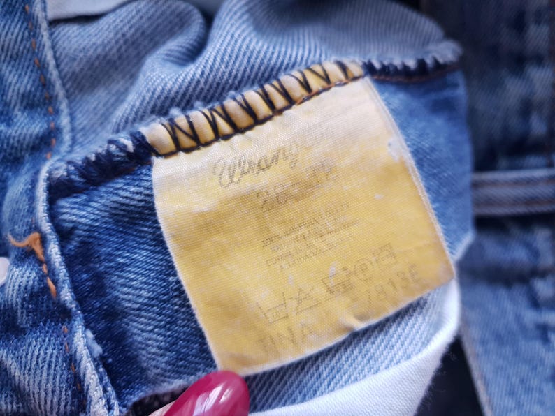 May include: Close-up of a pair of blue denim jeans. The yellowed tag reads "Wrangler" with size information and care instructions. The jeans show signs of wear.