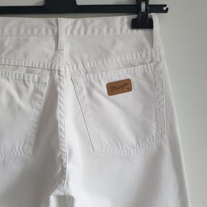 May include: Close-up of white denim jeans hanging on a black hanger. The jeans feature two back pockets and a brown leather patch with the "Wrangler" logo. The stitching and details of the pockets are visible.