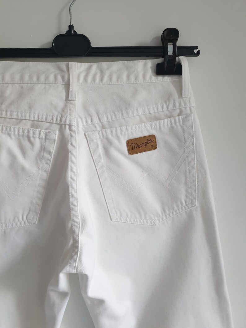 May include: Close-up of white denim jeans hanging on a black hanger. The jeans feature two back pockets and a brown leather patch with the "Wrangler" logo. The stitching and details of the pockets are visible.