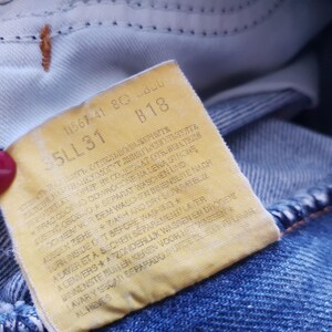 May include: Close-up of a faded yellow tag sewn into blue denim jeans. The tag features printed text, including the numbers "35LL31" and "B18". The jeans are a light blue wash, with visible stitching and a worn appearance.