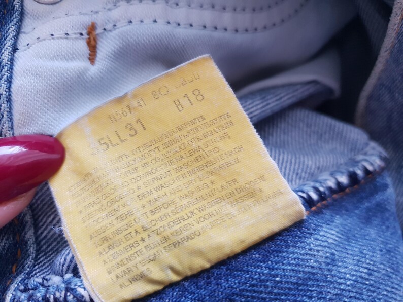 May include: Close-up of a faded yellow tag sewn into blue denim jeans. The tag features printed text, including the numbers "35LL31" and "B18". The jeans are a light blue wash, with visible stitching and a worn appearance.