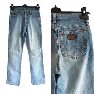 May include: Light blue denim jeans with a classic five-pocket design. The jeans feature a faded wash and a brown leather patch on the back pocket. The jeans are hanging on a black hanger.