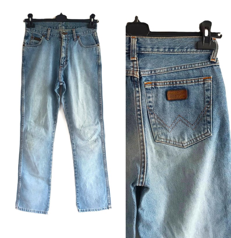 May include: Light blue denim jeans with a classic five-pocket design. The jeans feature a faded wash and a brown leather patch on the back pocket. The jeans are hanging on a black hanger.