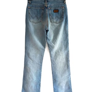 May include: Light blue denim jeans, viewed from the back, hanging on a black hanger. The jeans have a classic five-pocket design, with the Wrangler logo visible on the back pockets and a brown leather patch.