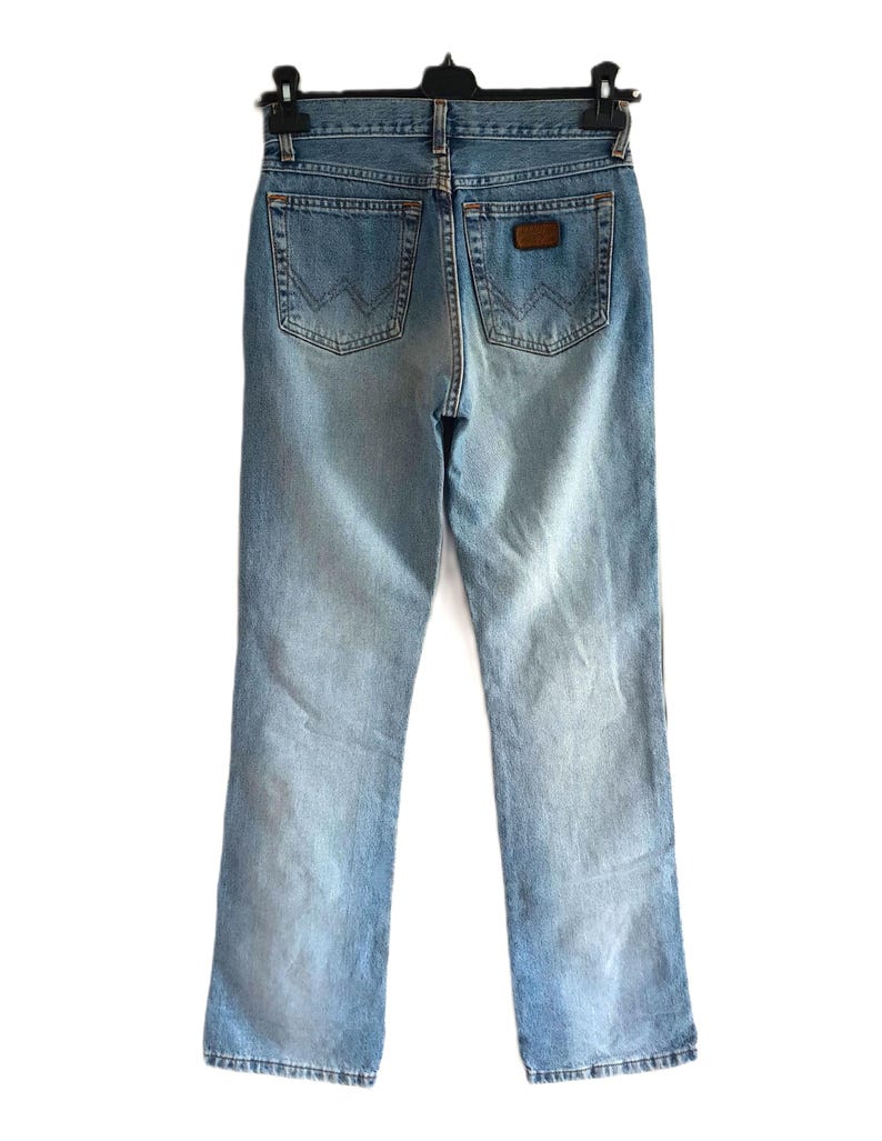May include: Light blue denim jeans, viewed from the back, hanging on a black hanger. The jeans have a classic five-pocket design, with the Wrangler logo visible on the back pockets and a brown leather patch.