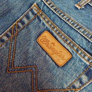 May include: Close-up of a blue denim pocket with a brown leather Wrangler label. The label features the Wrangler logo and a small horse silhouette.