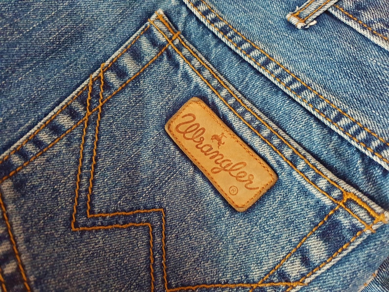 May include: Close-up of a blue denim pocket with a brown leather Wrangler label. The label features the Wrangler logo and a small horse silhouette.