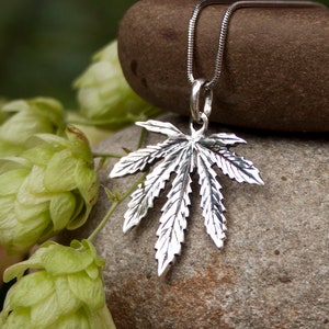 May include: A silver marijuana leaf pendant necklace on a silver chain, resting on a brown rock with green hops in the background.
