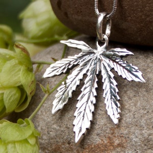 May include: A silver pendant in the shape of a marijuana leaf. The pendant is hanging from a silver chain and is resting on a rock.