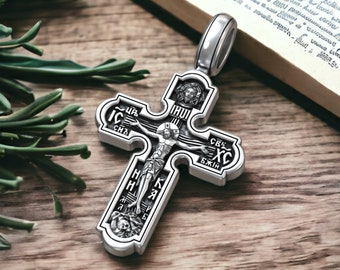 May include: A silver-colored Orthodox cross pendant with intricate detailing. The cross features a depiction of Jesus Christ and religious text. The pendant is displayed on a wooden surface with a book and greenery in the background.