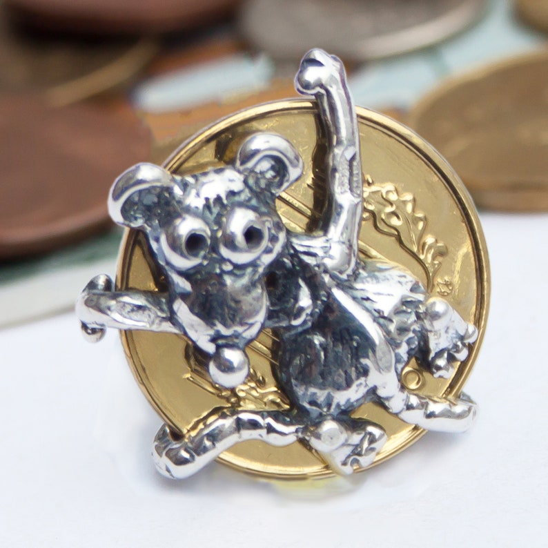 K&ouml;nnte beinhalten: Ein silberner Koalab&auml;ren-Anh&auml;nger mit strukturierter Oberfl&auml;che, der auf einer goldenen M&uuml;nze sitzt. Der Koala befindet sich in einer verspielten Pose mit einem erhobenen Arm.