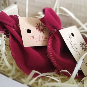 K&ouml;nnte beinhalten: Zwei rote Samtbeutel mit Kordelzug und braunen Papieretiketten mit der Aufschrift "Mia Silver Jewelry with meaning" und "Mia Silver Jewelry with meaning Studio".