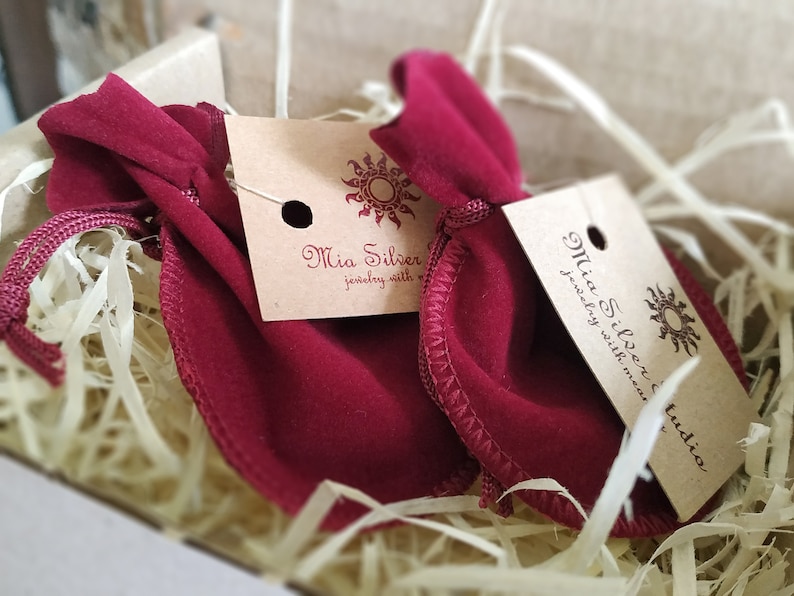 K&ouml;nnte beinhalten: Zwei rote Samtbeutel mit Kordelzug und braunen Papieretiketten mit der Aufschrift "Mia Silver Jewelry with meaning" und "Mia Silver Jewelry with meaning Studio".