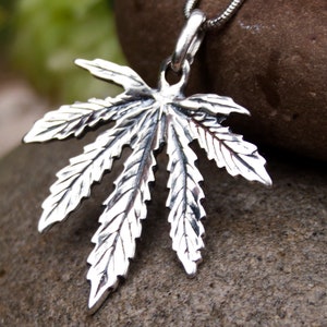 May include: A silver pendant in the shape of a marijuana leaf. The pendant is detailed with realistic veins and a textured surface.