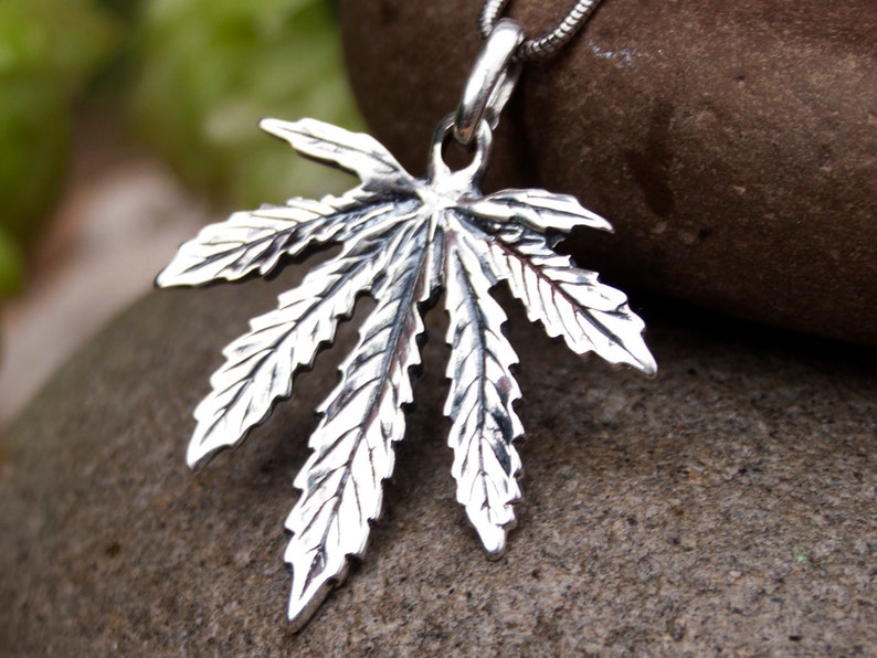 May include: A silver pendant in the shape of a marijuana leaf. The pendant is detailed with realistic veins and a textured surface.