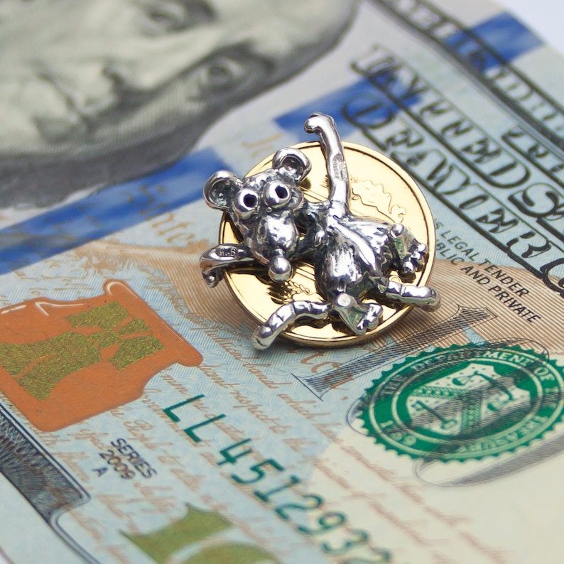 K&ouml;nnte beinhalten: Ein silberner Anh&auml;nger mit einem Affen, der auf einer goldenen M&uuml;nze sitzt. Die M&uuml;nze tr&auml;gt die Aufschrift "United States of America" und "One Dollar". Der Anh&auml;nger liegt auf einem US-Dollar-Schein mit der Aufschrift "Legal Tender" und "Public and Private".