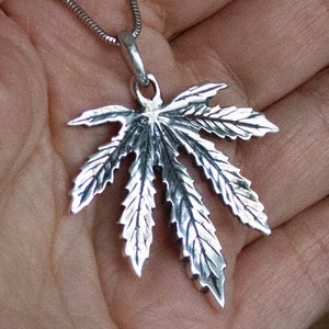 May include: A silver pendant in the shape of a marijuana leaf. The pendant is detailed with veins and serrated edges.