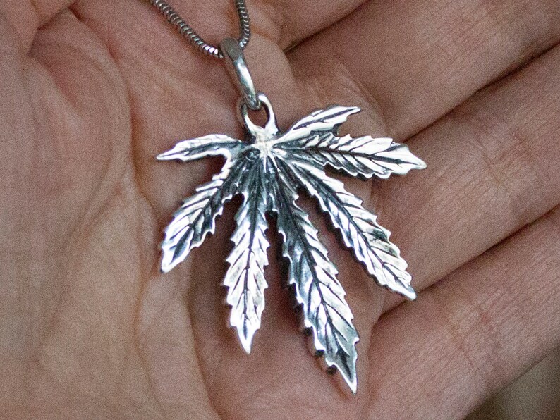 May include: A silver pendant in the shape of a marijuana leaf. The pendant is detailed with veins and serrated edges.