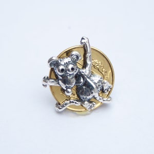 K&ouml;nnte beinhalten: Ein silberner Koalab&auml;ren-Anh&auml;nger mit einem goldenen M&uuml;nzenhintergrund. Der Koala ist in einer verspielten Pose mit erhobenen Armen.