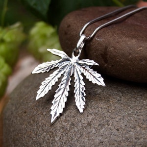 May include: A silver cannabis leaf pendant necklace on a silver chain. The pendant is detailed with realistic leaf veins and is hanging from a smooth, gray stone.
