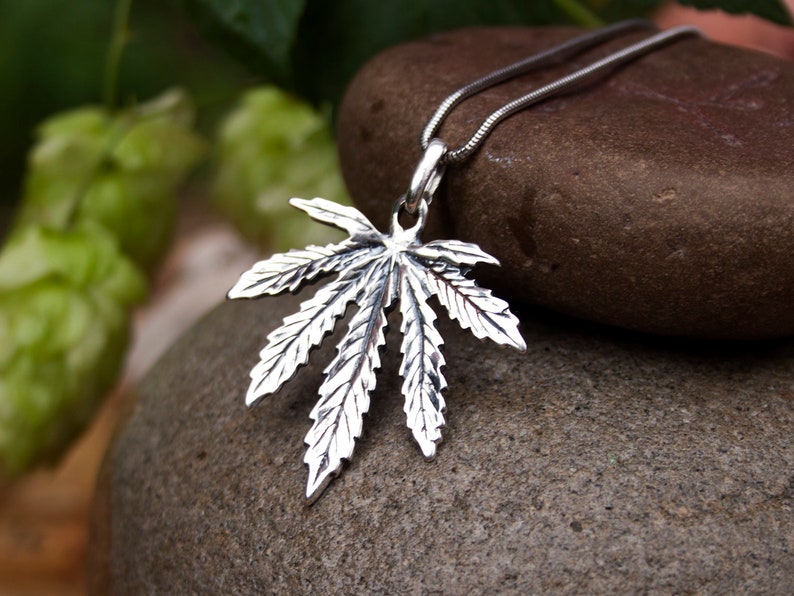 May include: A silver cannabis leaf pendant necklace on a silver chain. The pendant is detailed with realistic leaf veins and is hanging from a smooth, gray stone.