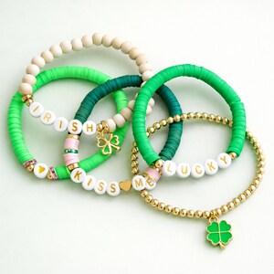 May include: A collection of beaded bracelets in shades of green, white, and gold. Some bracelets feature the words "IRISH", "KISS ME", and "LUCKY" spelled out with white letter beads. Charms include a gold shamrock and heart.