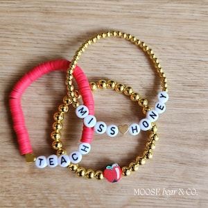 May include: Three bracelets with gold beads and white letter beads spelling "Teacher", "Miss", and "Honey". The "Miss" bracelet has a small gold heart charm and the "Honey" bracelet has a red apple charm.