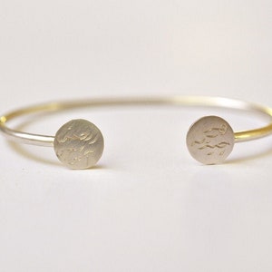 May include: A silver-colored cuff bracelet with a minimalist design. The bracelet features two circular end pieces with etched patterns. The open design allows for easy wear. The jewelry is suitable for everyday wear.