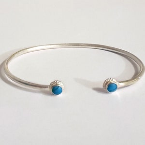 May include: A silver-colored cuff bracelet with a turquoise stone at each end. The bracelet has a simple, open design. The turquoise stones are round and set in a textured, circular setting.