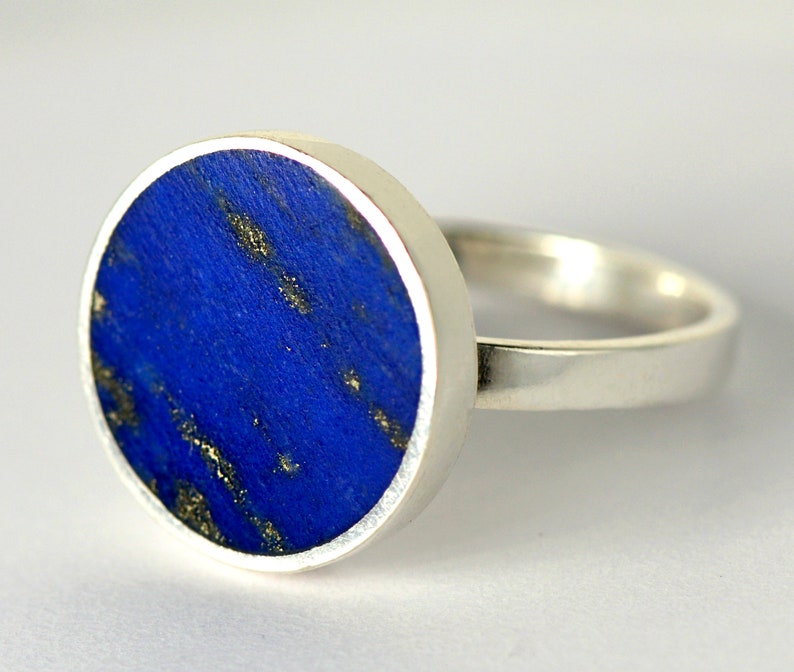 May include: A silver ring with a round blue lapis lazuli stone set in the center. The stone has a marbled pattern with gold flecks.
