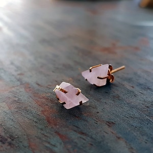 Könnte beinhalten: Zwei rosafarbene Ohrstecker aus Rosenquarz mit goldfarbenen Metallakzenten. Die Ohrringe haben einen rohen, natürlichen Look, wobei die rosa Quarzsteine in eine dreieckige Form geschnitten sind. Die Stecker sind vor einem blau-grünen Hintergrund platziert.