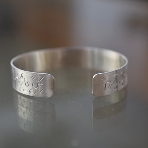 May include: A silver cuff bracelet with etched designs. The open-ended bracelet is made of a shiny, metallic material. The etched designs appear to be stylized text or symbols. The bracelet is displayed on a reflective surface.