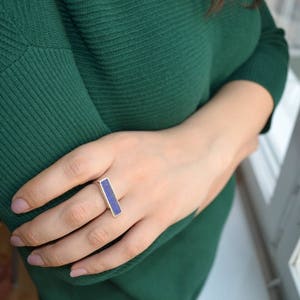 以下が含まれることがあります： 長方形の濃い青色の石が付いたシルバーリング。 リングは指に着用され、手は濃い緑色の長袖トップスに寄りかかっています。 背景はぼやけており、屋内環境を示唆しています。
