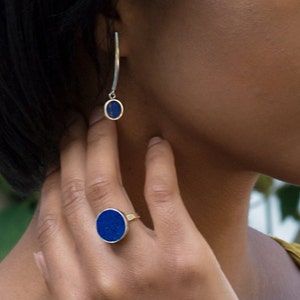 May include: A silver ring with a blue oval lapis lazuli stone. The ring is worn on the index finger of a person's hand. The person is also wearing a silver earring with a blue oval lapis lazuli stone.