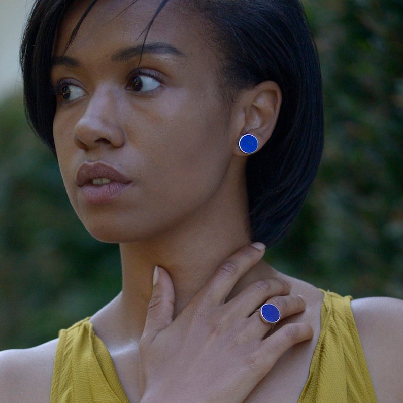 May include: A woman wearing a yellow top with a blue ring and matching blue earrings. The ring and earrings are round and have a silver setting.
