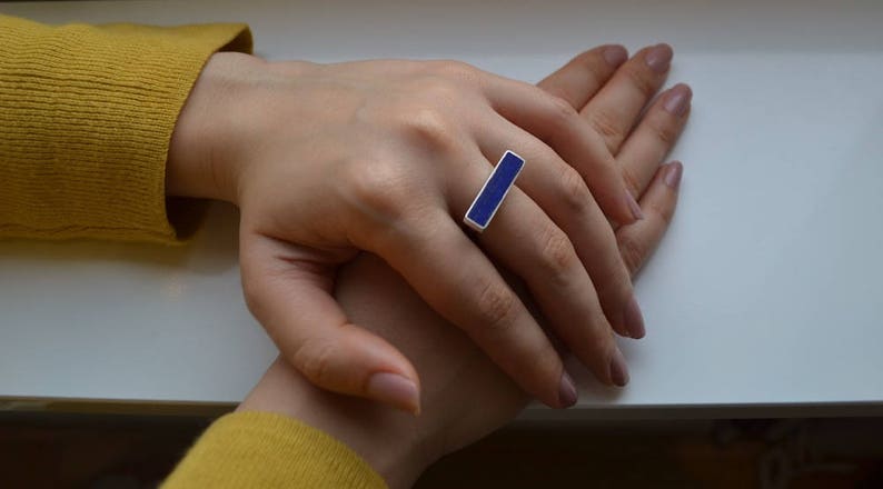 以下が含まれることがあります： 長方形の濃い青色のエナメル象嵌が施されたシルバーリング。リングは指に着用され、手は白い表面に置かれています。その人は黄色のセーターを着ています。