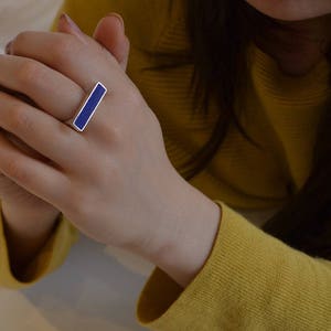 以下が含まれることがあります： 長方形の濃い青色のインレイが付いたシルバーリング。リングは指に着用されており、手と腕が見えます。その人は黄色のセーターを着ています。リングはシンプルでモダンなデザインです。