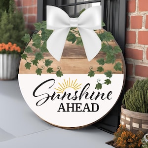 Puede incluir: Un letrero redondo de madera con un lazo blanco, hojas verdes y el texto "Sunshine Ahead" en negro con un gráfico de sol dorado.