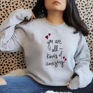 Könnte beinhalten: Ein grauer Pullover mit dem Text "you are -all- kinds of amazing" in schwarzer Schrift und kleinen roten Herzen.