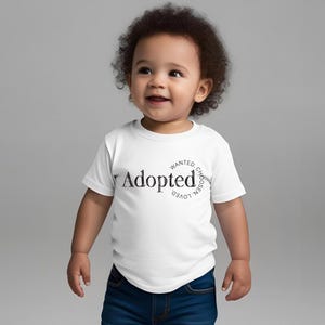 May include: A white t-shirt featuring the word "Adopted" in a decorative font, with the words "Wanted, Loved, Chosen" arranged in a circle. The shirt is worn by a smiling toddler in blue jeans.