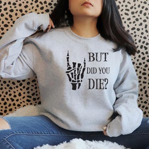 Puede incluir: Una sudadera gris con una mano de esqueleto negra haciendo un signo de rock and roll y el texto "BUT DID YOU DIE?"