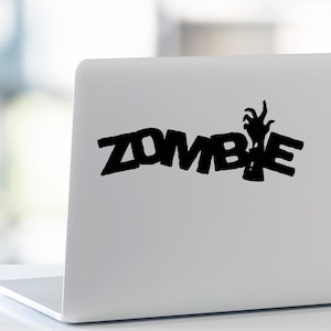 Puede incluir: Pegatina negra con la palabra "ZOMBIE" en letras negritas y estilizadas. La letra "E" se sustituye por un gráfico de una mano de zombi extendida. La pegatina está en un portátil blanco.