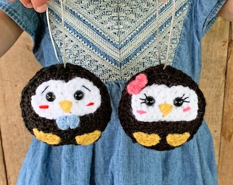 Könnte beinhalten: Zwei gehäkelte Pinguin-Anhänger, einer mit einer blauen Schleife und der andere mit einer rosa Schleife. Beide Pinguine haben einen schwarz-weißen Körper mit gelben Füßen und schwarzen Augen.